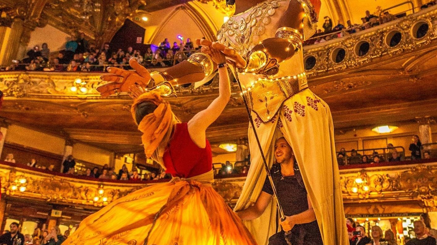 Giant puppet dancing with performer on a stage, audience watching.