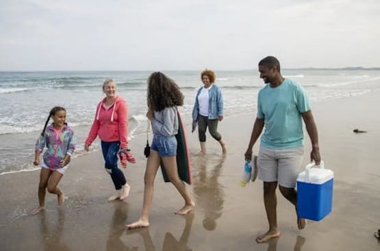 Family walking along the beach, carrying cooler and surfboard.