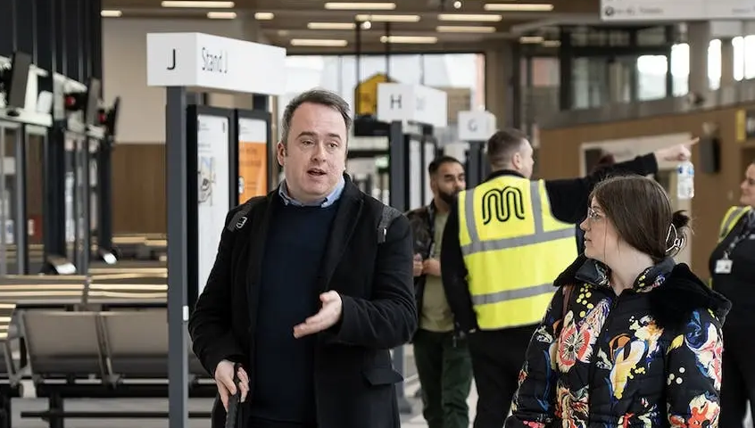 A man with a white cane is talking in a brightly lit building with other people.