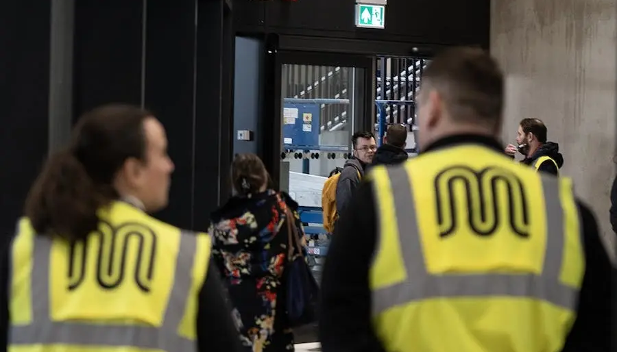 People walking in a building; security guards in vests with a logo.