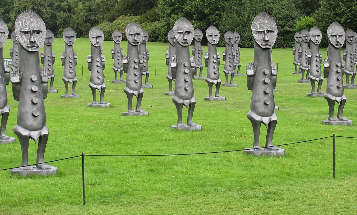 Statues in a field. Grey statues with human faces, hands up. Green grass background.