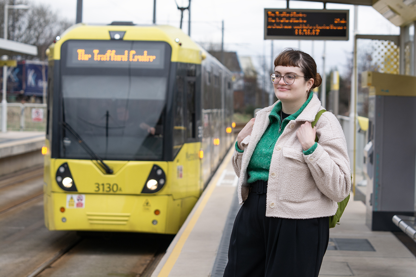 Woman by a tram at a station. The tram's destination is "The Trafford Centre."