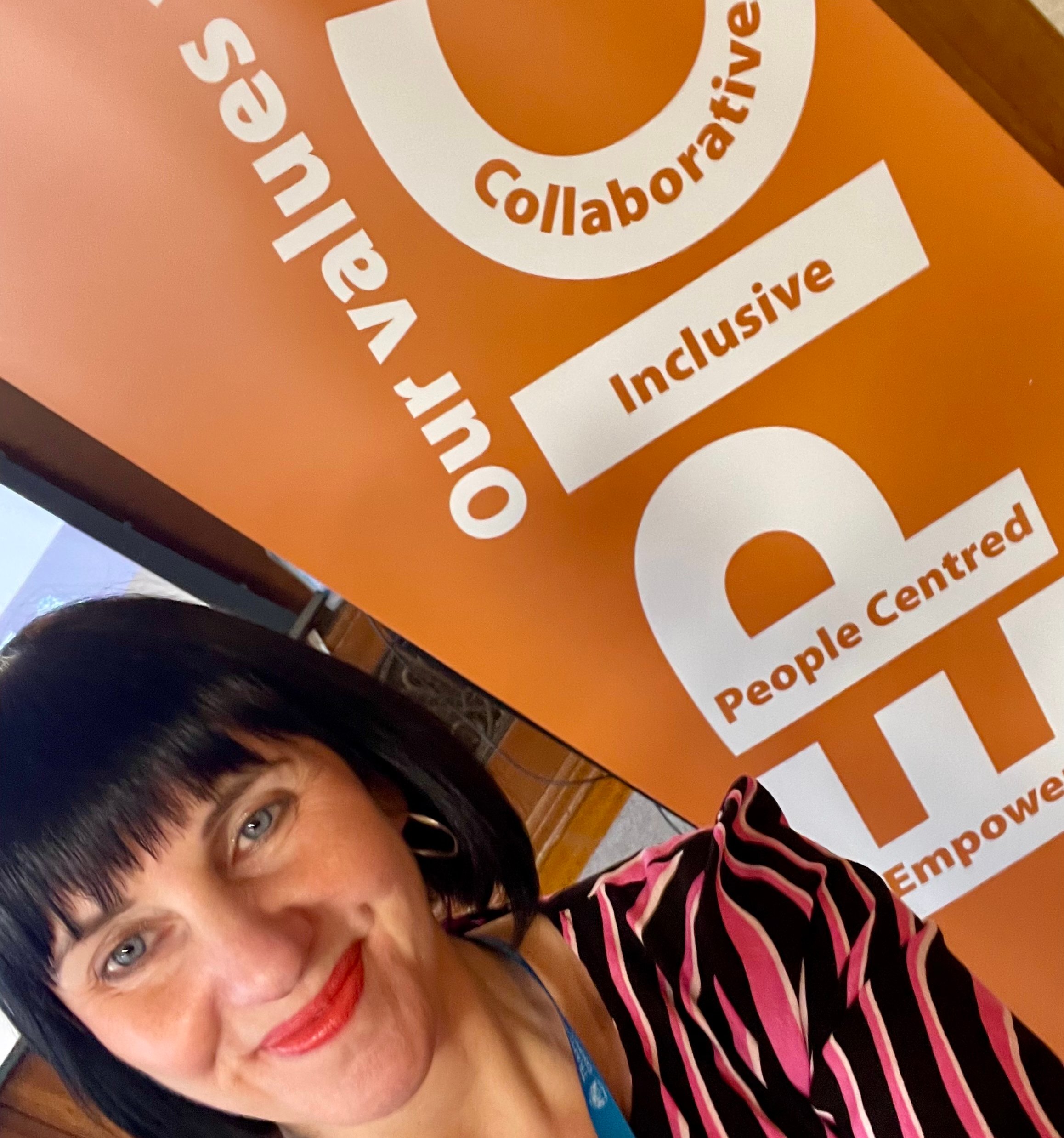 Woman with black hair smiling, Trafford Council lanyard, orange background with words.