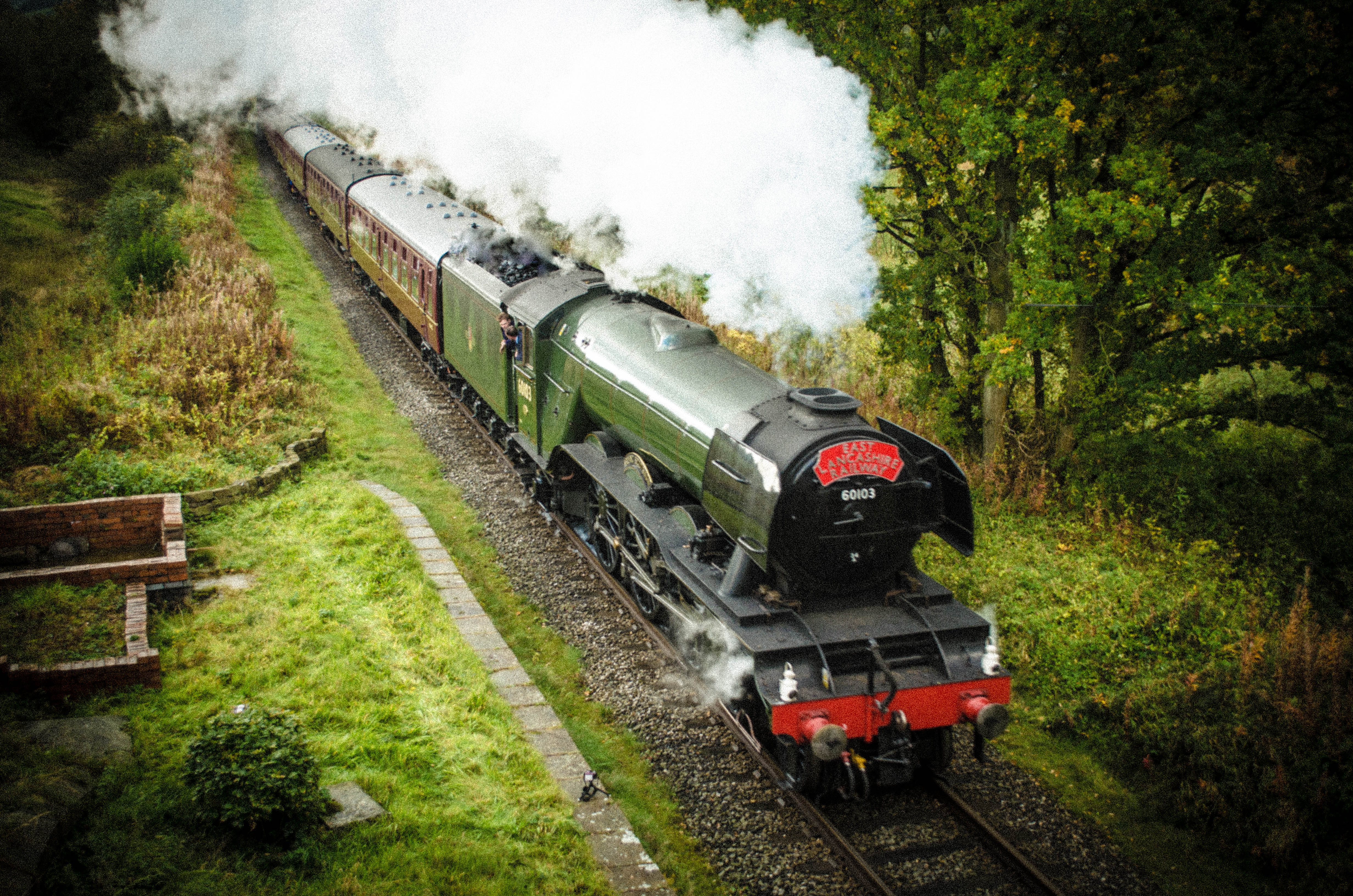 Steam train on tracks by a green field, trees, and stone wall.