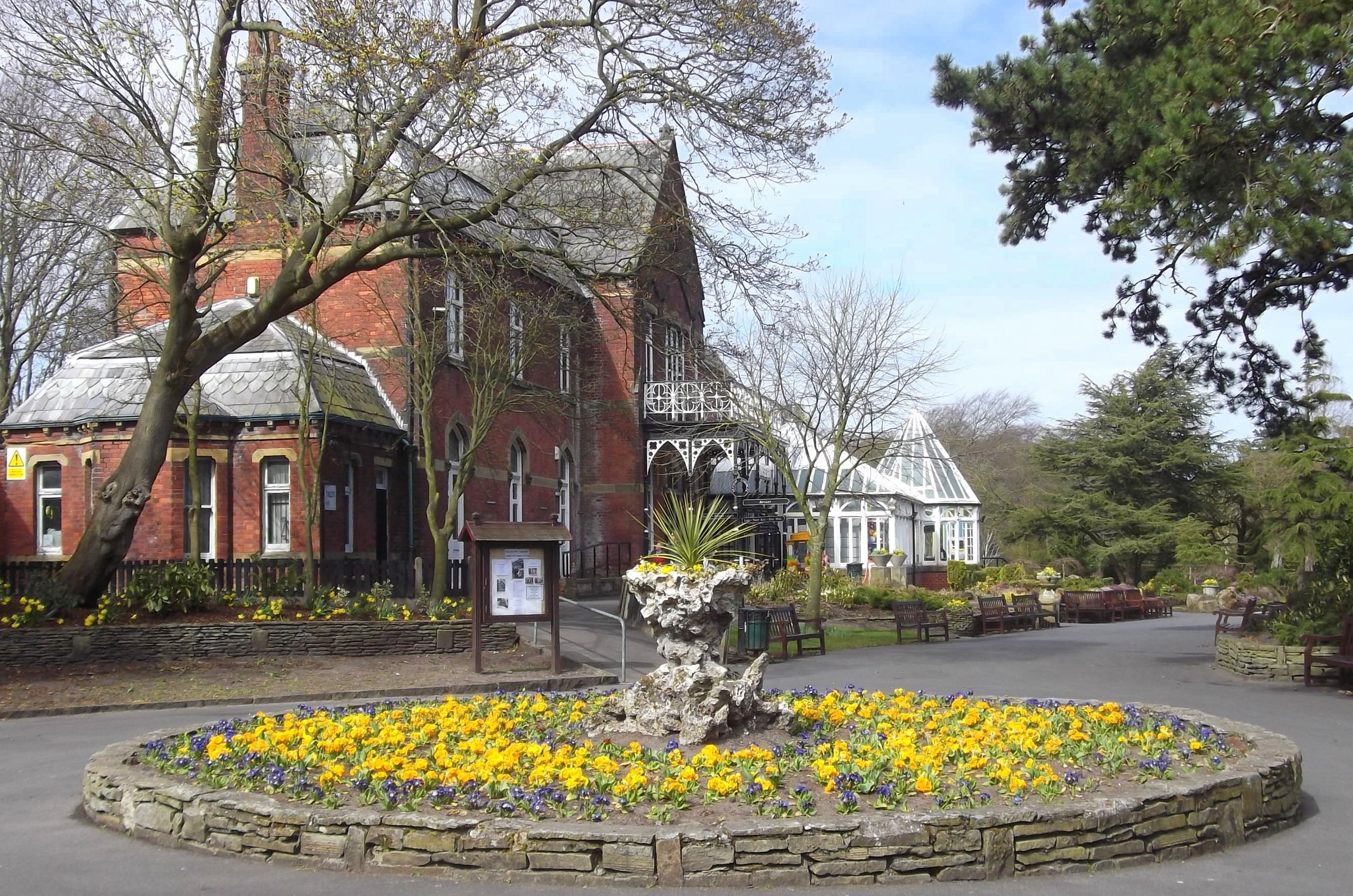 Gardens with flower beds, brick building, trees, and benches.