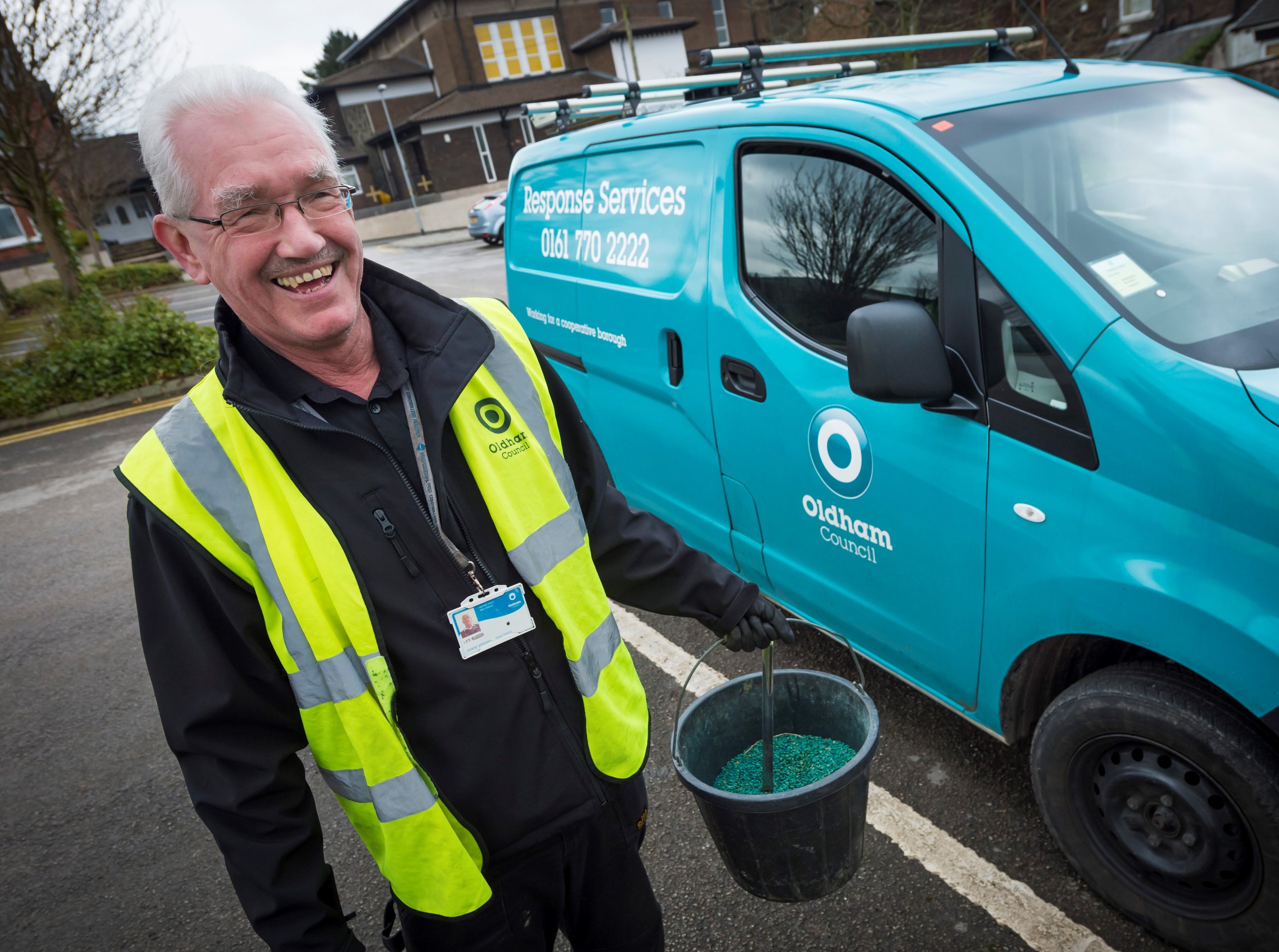 Man holding a bucket, standing next to a teal van, wearing a safety vest.