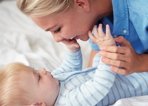 Woman smiles at a baby, touching its hand. Baby is on a bed.