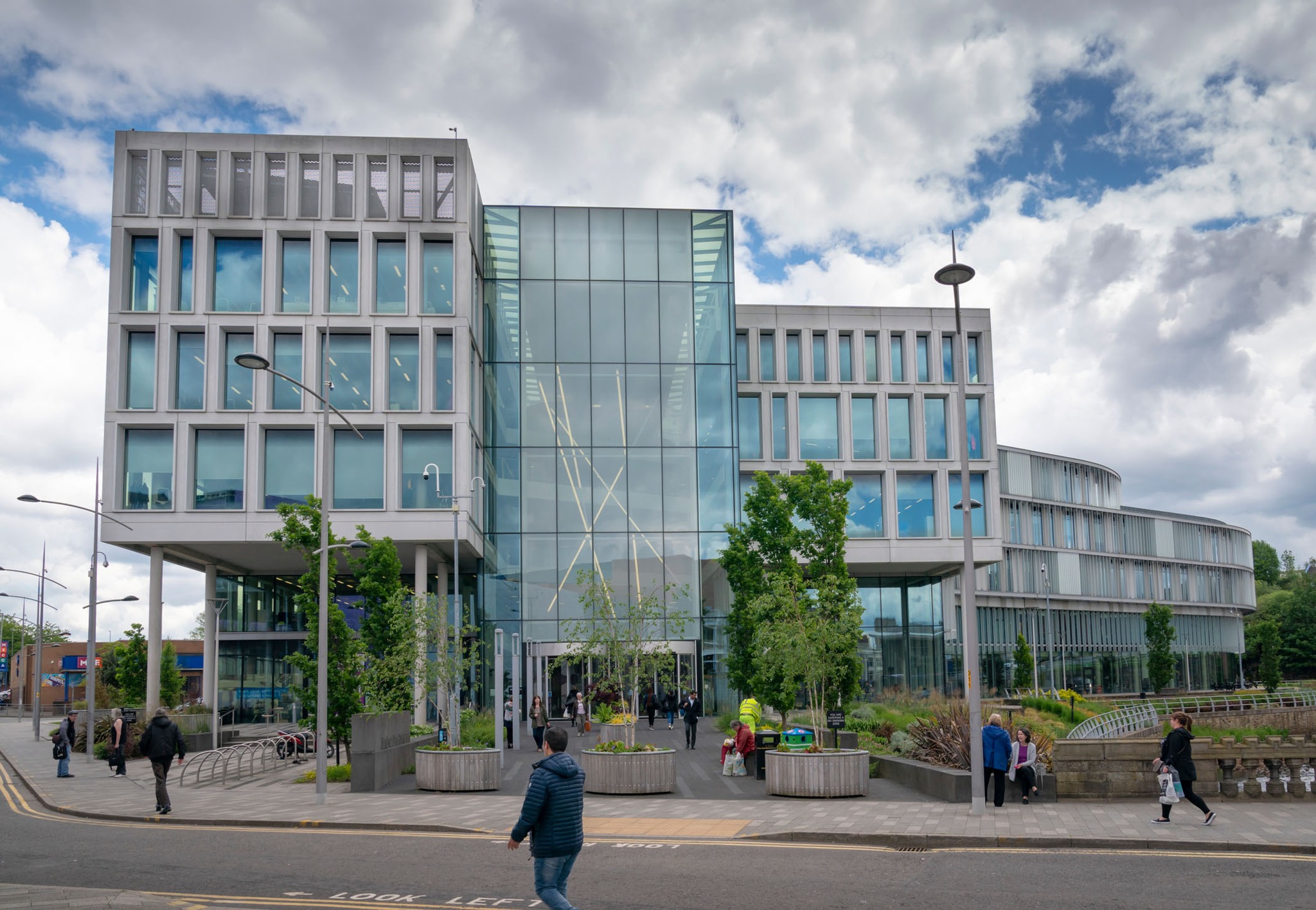 Modern building with glass facade, trees, pedestrians, and cloudy sky.