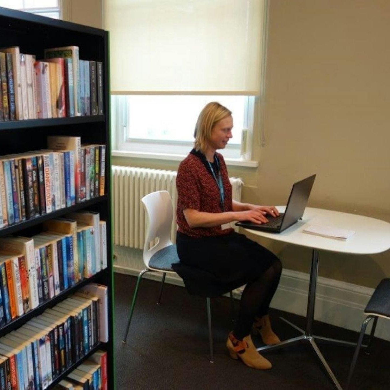 A woman using a laptop next to a bookshelf in a library setting.