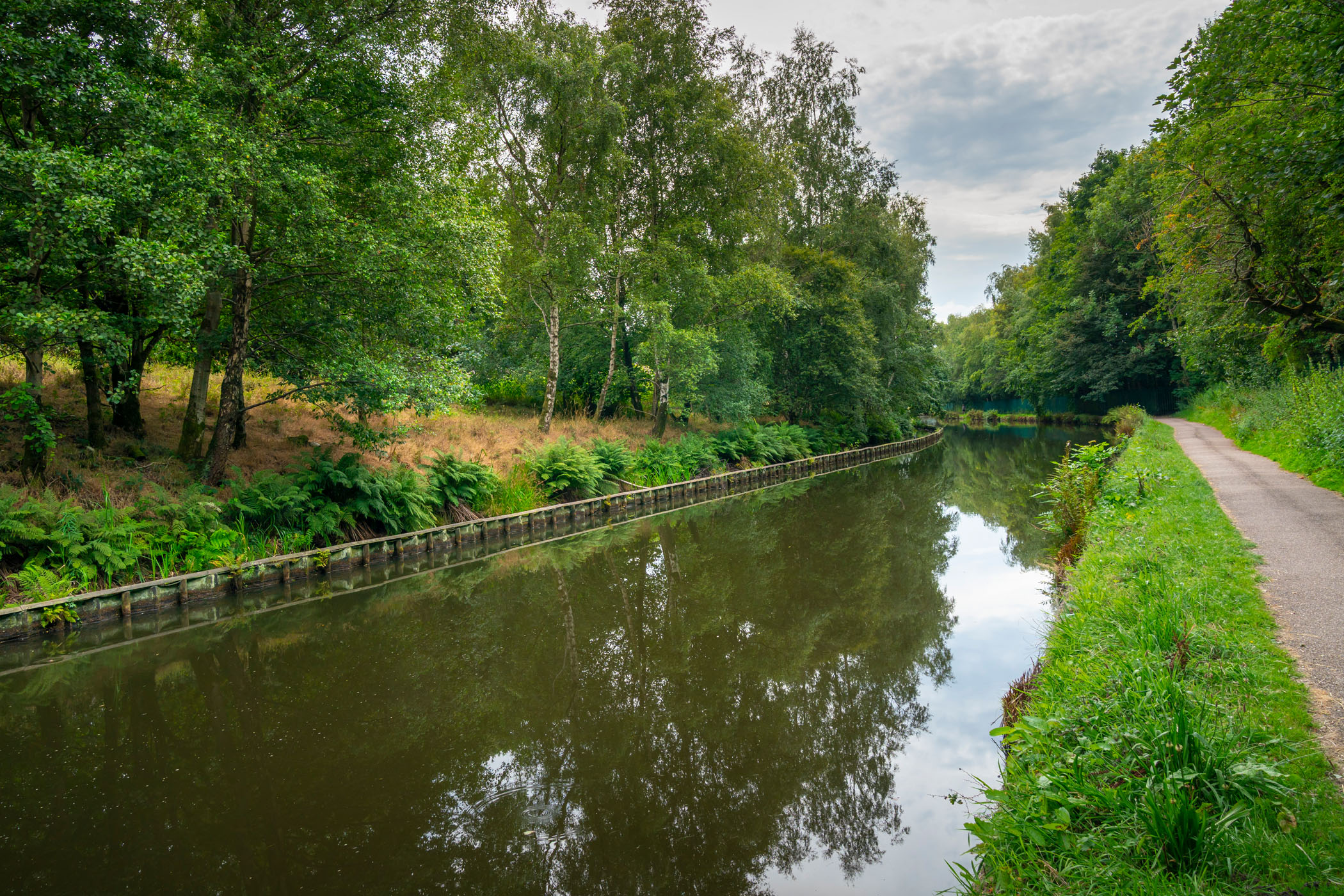 Canal with green trees and a path along the side. The water reflects the trees and the sky.
