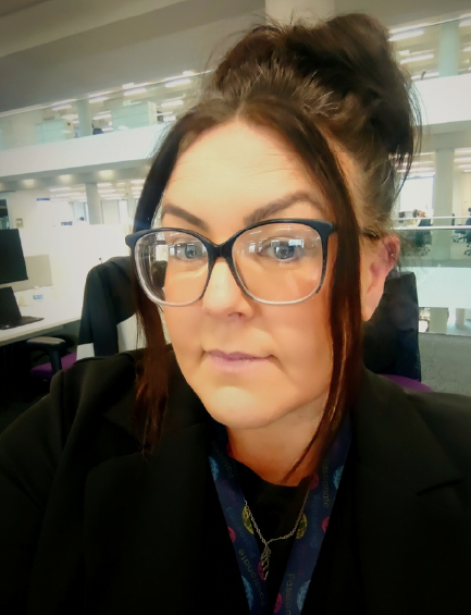 A woman with dark hair pulled up in a bun, wearing glasses and a black blazer, smiles for a selfie in an office environment.