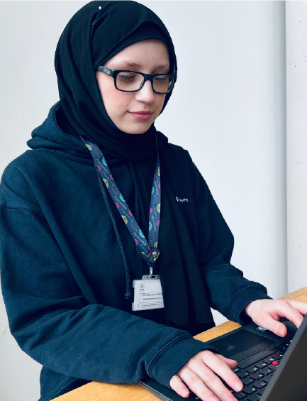 Young woman wearing glasses and a black hijab, typing on a laptop with a lanyard around her neck.