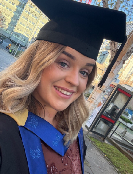 A woman with blonde hair smiles for a selfie in her graduation cap and gown, outdoors.