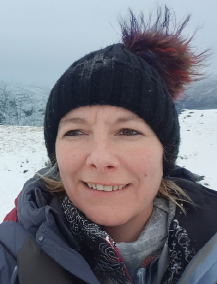 Woman in a black beanie with a colorful pompom smiles during a snowy hike, wearing a grey jacket and patterned scarf.