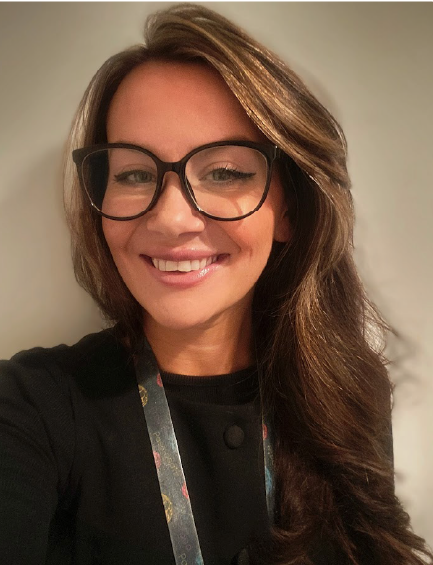 A woman with brown wavy hair and glasses smiles, wearing a black sweater and a lanyard.