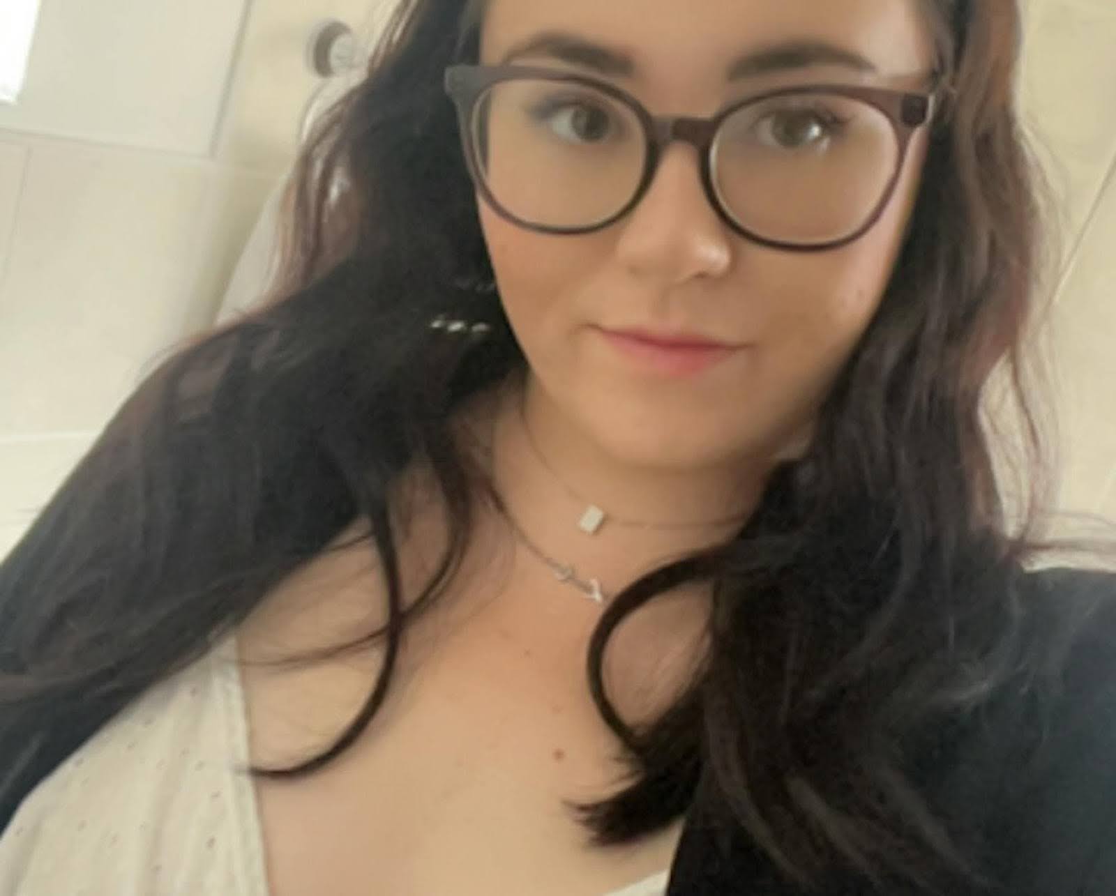 A young woman with dark hair and glasses smiles while wearing a white top and a dark jacket.