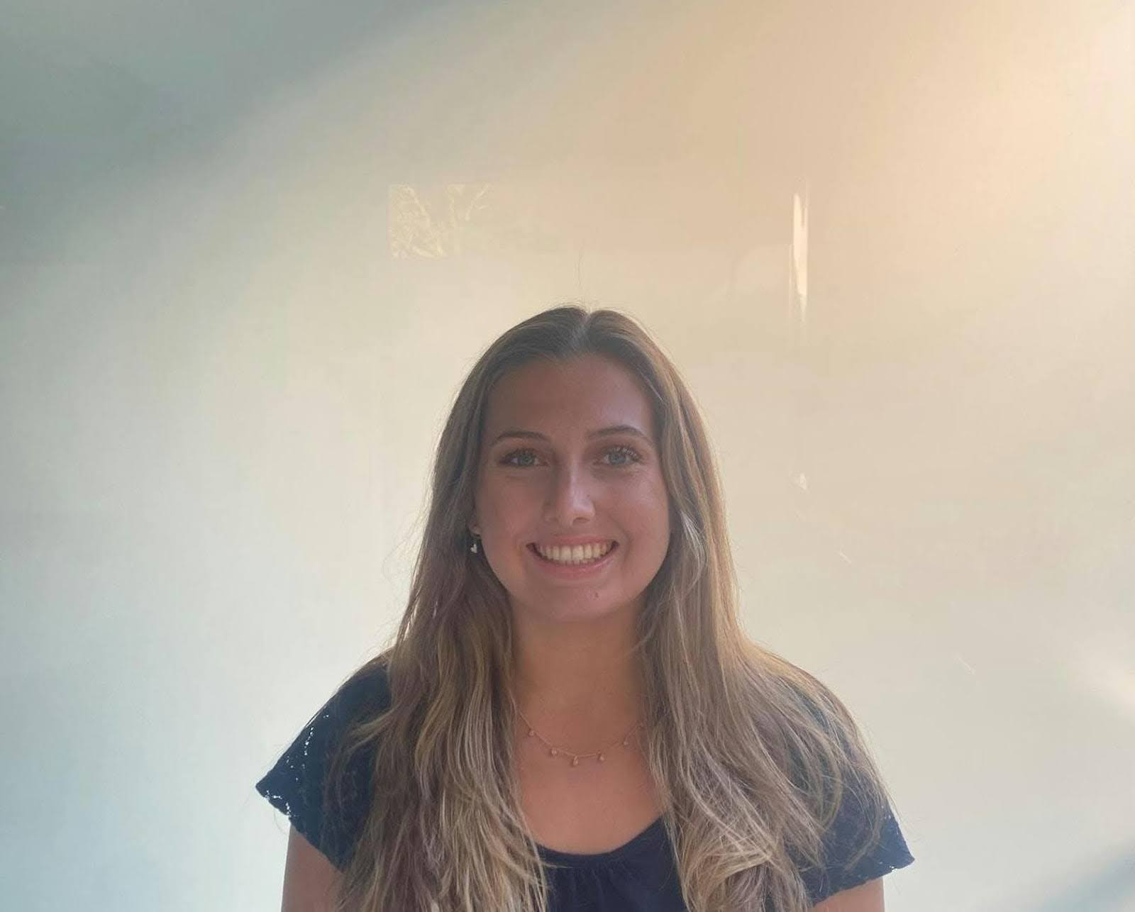 A woman with long, blonde hair and blue eyes smiles warmly. She is wearing a dark blue shirt and a delicate necklace.