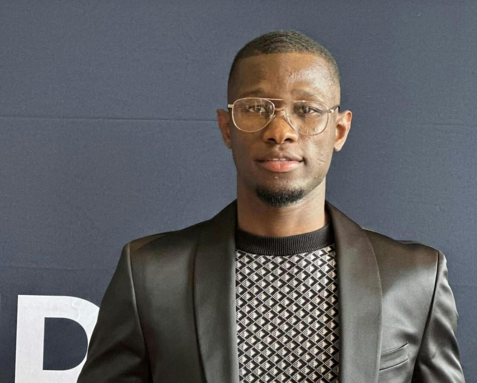 A young Black man wearing glasses, a black suit jacket, and a patterned sweater stands against a dark blue backdrop.