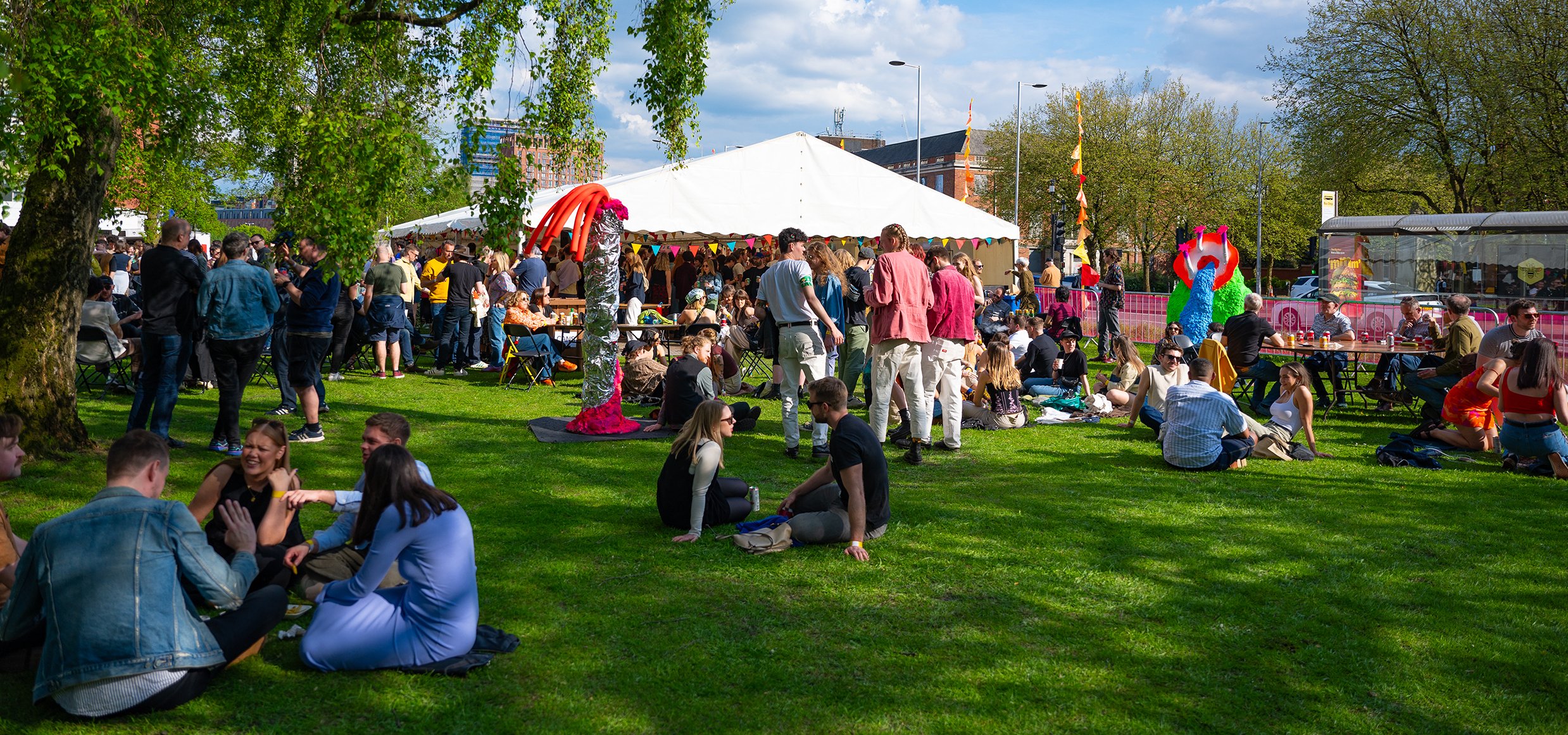 People relax on the grass at an outdoor festival with a large white tent, colorful flags, and art installations.