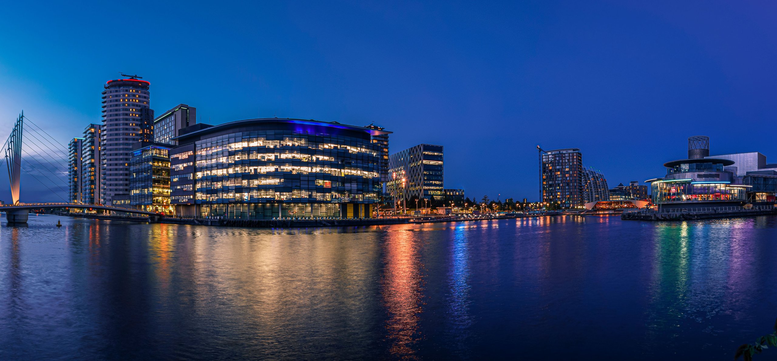 Panoramic view of MediaCityUK waterfront at dusk with illuminated buildings reflecting in the water.